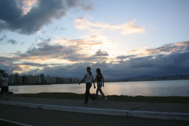 Pessoas caminhando pela orla de Florianópolis durante o pôr do sol, com prédios e montanhas ao fundo.