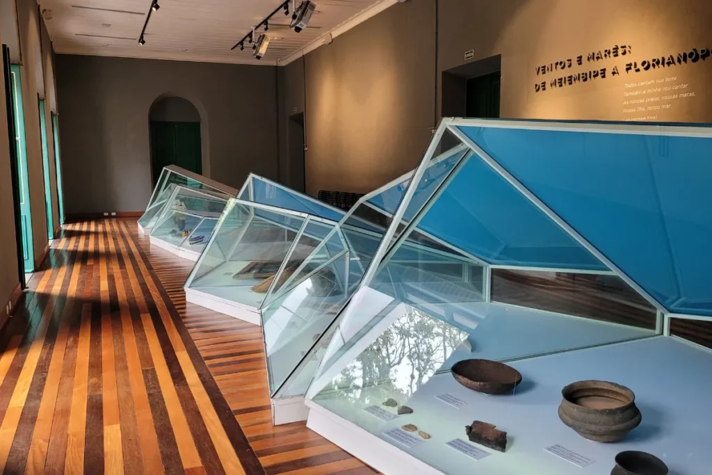 Sala de exposição do Museu de Florianópolis com vitrines, objetos históricos e piso de madeira.