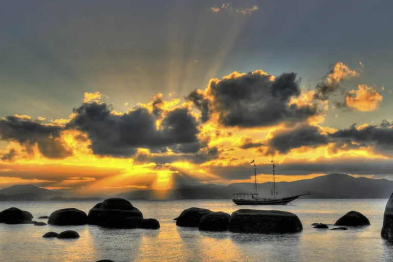 Pôr do sol em Santo Antônio de Lisboa com barco, pedras no mar e montanhas ao fundo.