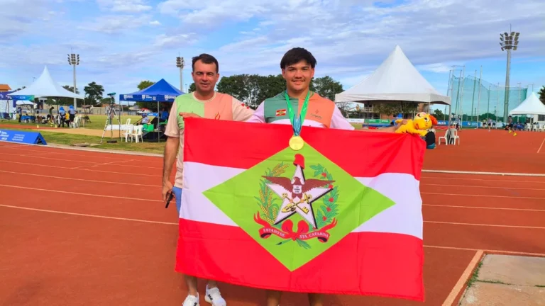 Atleta catarinense com medalha de ouro no peito segura a bandeira de Santa Catarina ao lado do técnico na pista de atletismo