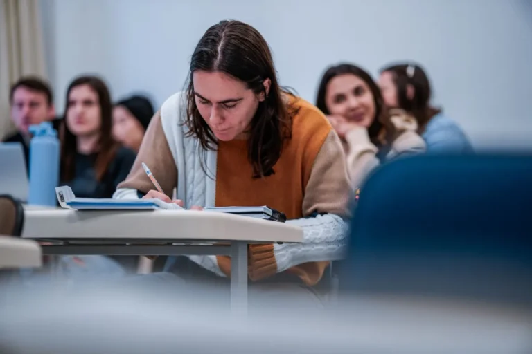 Estudante universitária escreve no caderno durante aula, com colegas ao fundo em sala de faculdade de Santa Catarina